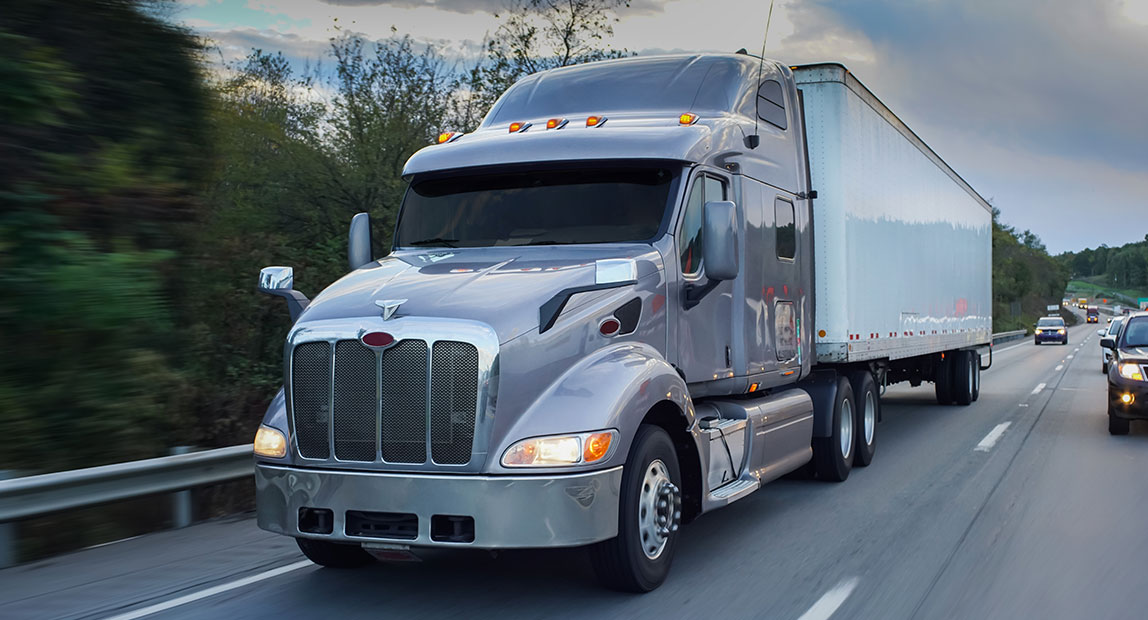 interior-trucking-md A silver semi-truck driving on a highway with other vehicles in the background and trees lining the road.